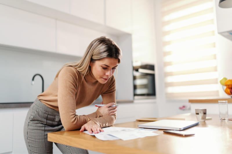 VA Loan Refinance Options 1 A woman stands in a modern kitchen, leaning over a wooden counter while reviewing documents. A tablet, glass of water, and bowl of fruit are also on the counter beside her.