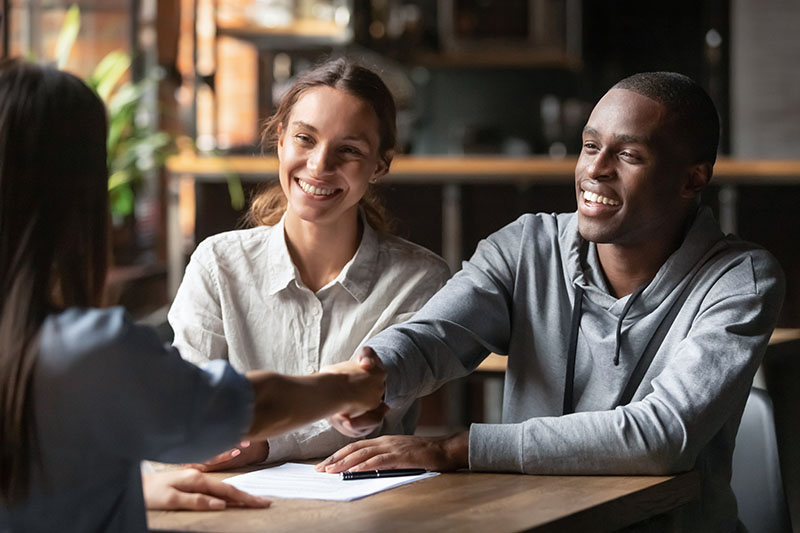 VA Home Loan 1 A smiling man and woman sit at a table, shaking hands with a third person whose back is to the camera. Papers and a pen are on the table, suggesting a meeting or agreement in a bright, casual setting.