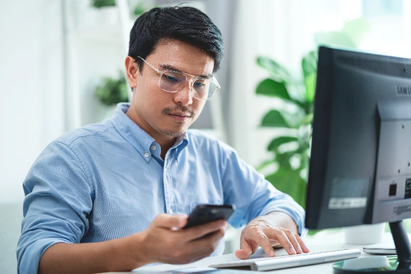Blog 3 A man wearing glasses is sitting at a desk with a computer monitor and a cell phone